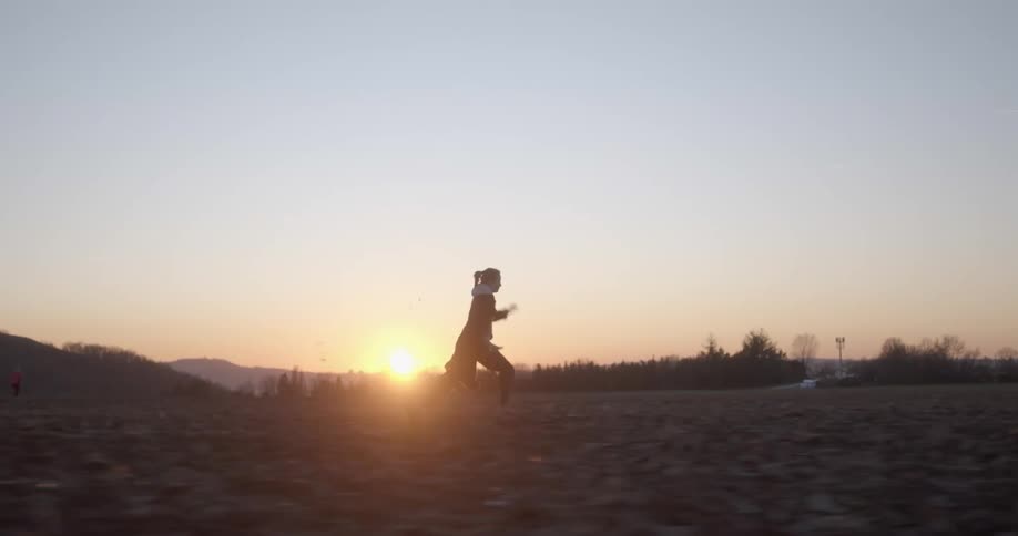 A person running on a grassy field with trees the sunset the sky and clouds in the background