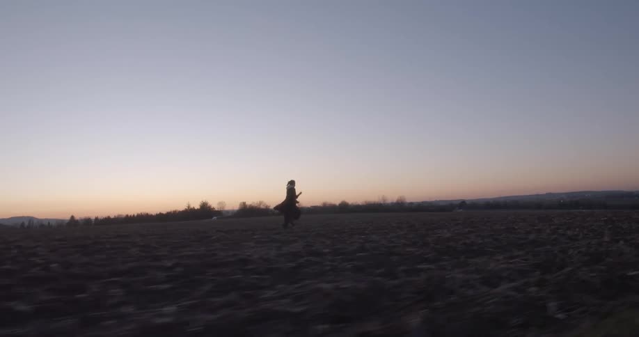 Young woman wearing a coat running in a farm field stops running to throw a rock at evening
