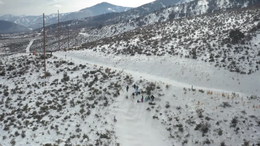 People sliding down a snowy slope with sleds with a mountain range the sky and clouds in the background
