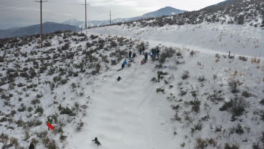 A group of people holding sleds and toboggans watch as someone slides down the snow covered slope