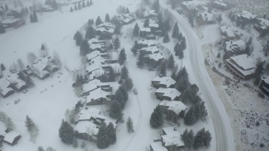 An aerial view of a neighborhood covered in snow with a car driving down a road with hills in the background