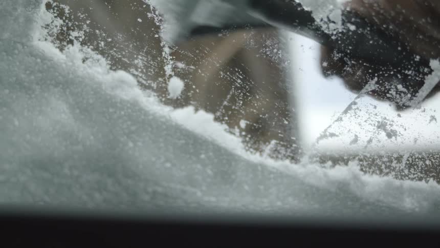 A person is using a scraper to clear away snow from the windshield of a vehicle