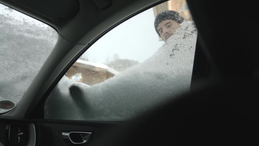 A man wiping snow and ice from a car window on a cold winter day