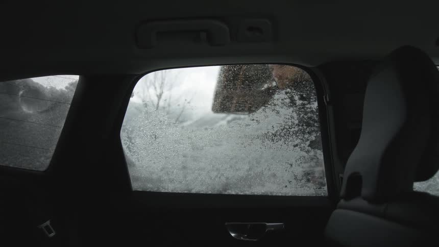 A man removing snow from a vehicles window with the sky and clouds in the background