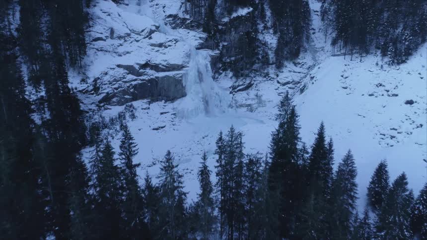 A waterfall spills down a snowy cliff in an evergreen forest during the evening