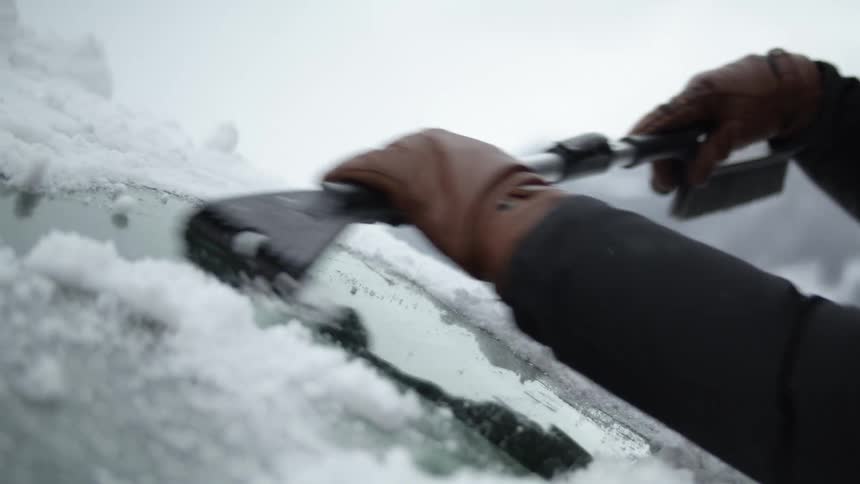 A person wearing brown leather gloves scrapes frost and ice from a winshield