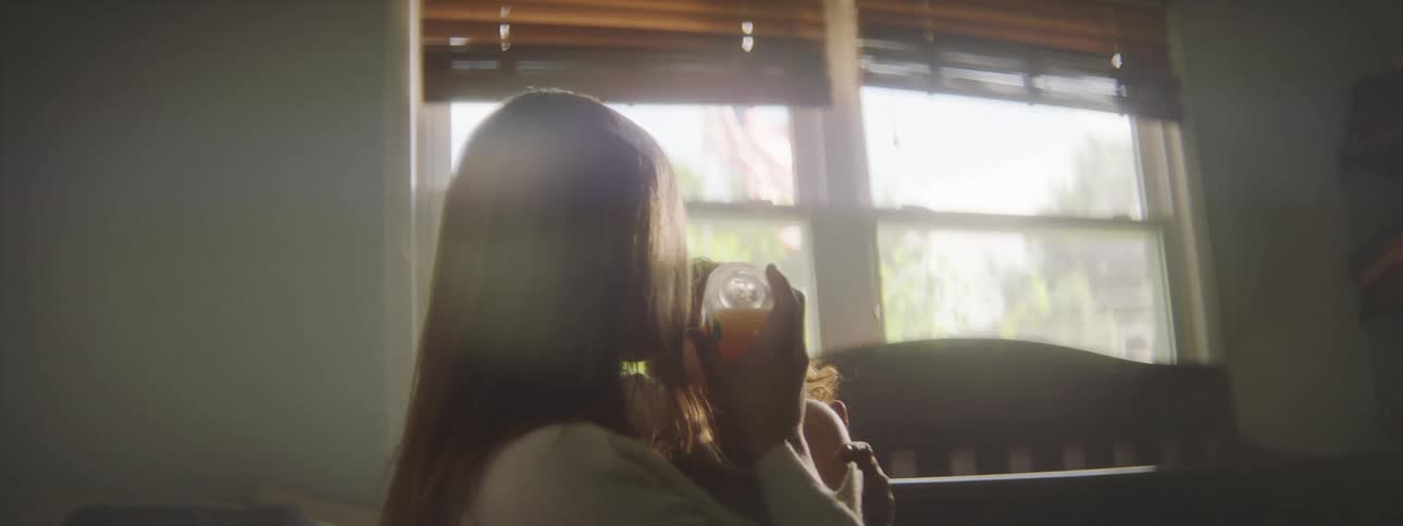 A woman in a room feeding a baby with a bottle with a crib and window in the background