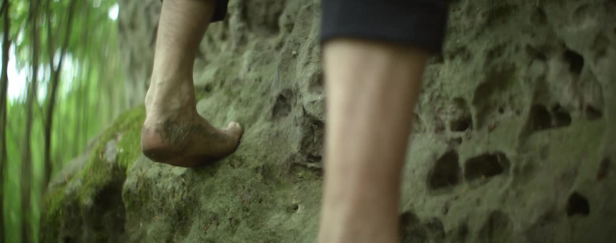 A barefoot man climbing the rugged face of a rock wall in the forest
