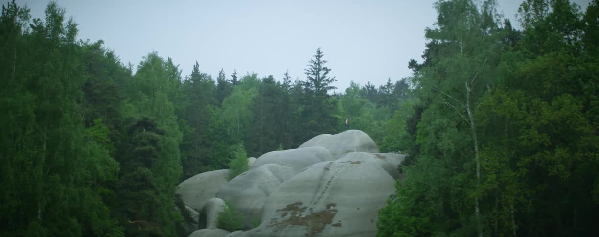 A person walking on a cliff surrounded by trees with the sky and clouds in the background