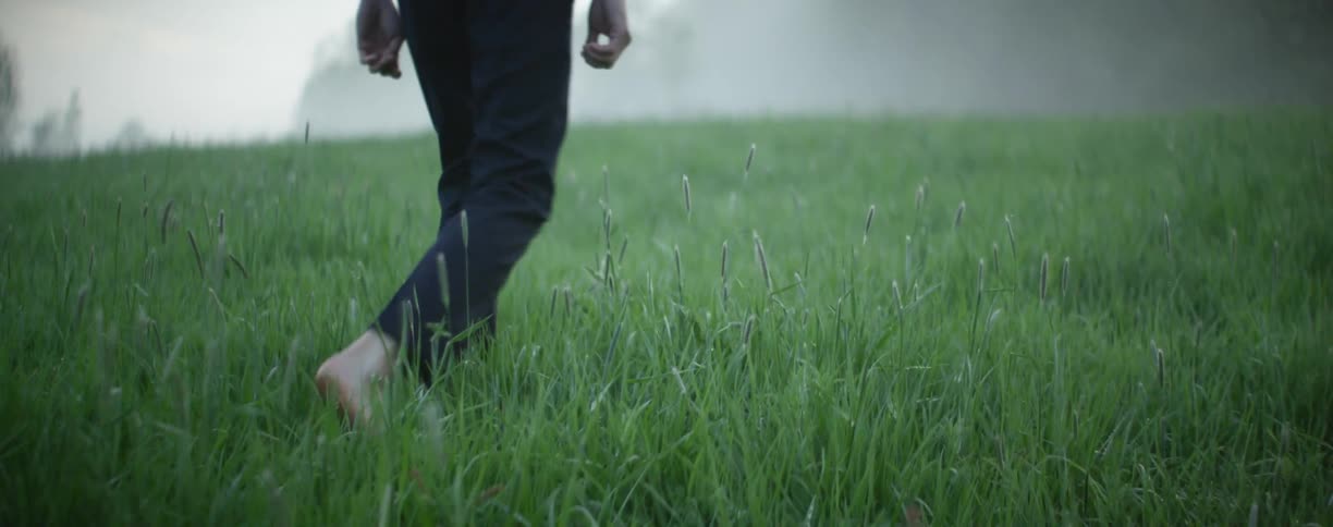 A man walks barefoot through a grassy field with fog in the distance