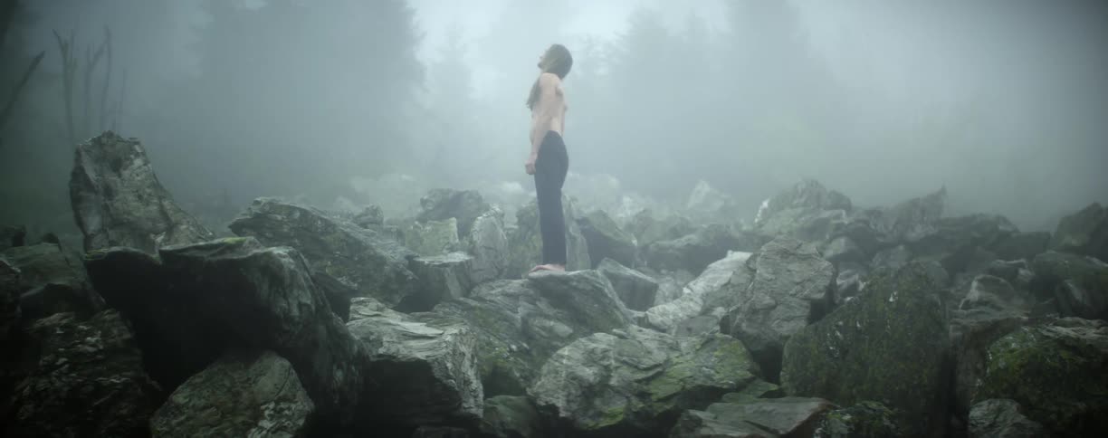 A man stands on a large pile of rocks and looks up at the foggy sky