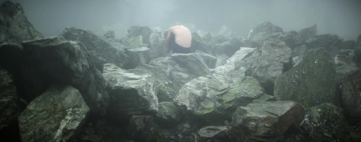 A man cradles his knees while sitting atop a pile of large stones on a misty day