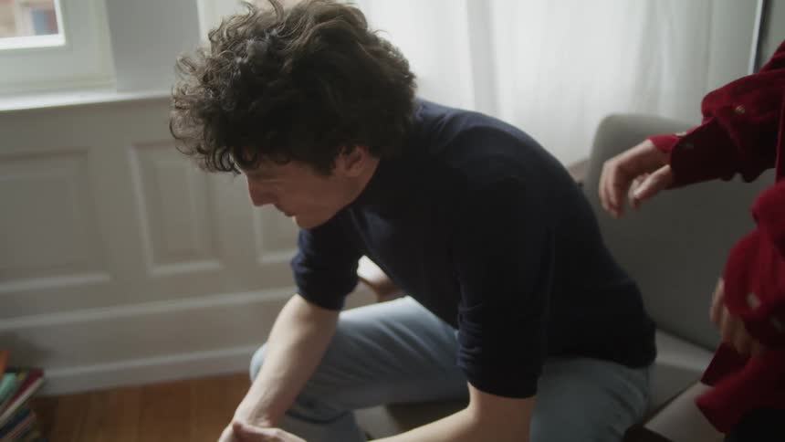 A man in a room sitting on a chair as another person touches his shoulder with a wall and window in the background