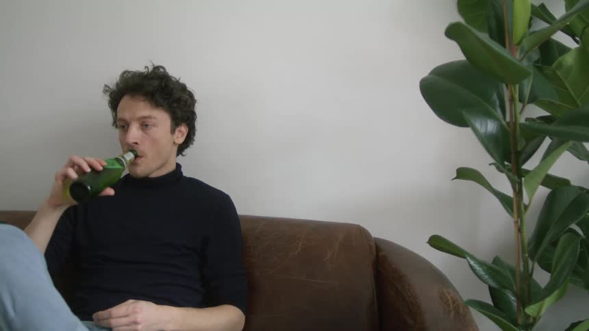 Man sitting on a brown leather couch in the living room at home drinks beer while talking
