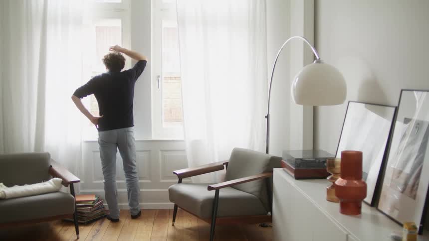 A man walks across a room in a house to gaze out of a window while leaning against the window frame