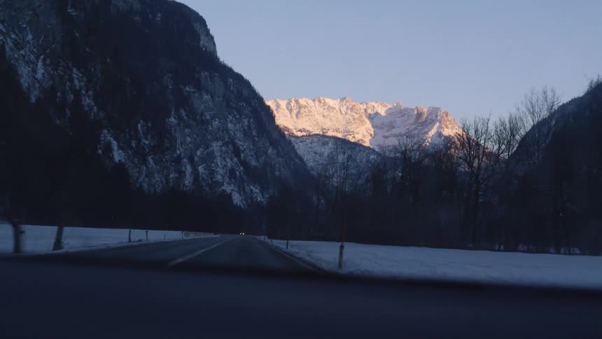 A view of a car driving down a road at the base of a mountain