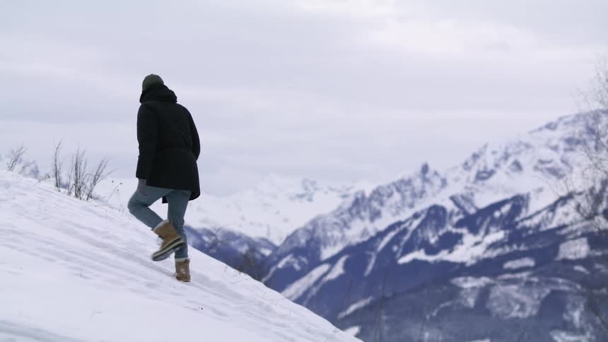 A man walks up a snowy mountain slope while looking out at the distant mountains and landscape below on a cloudy day