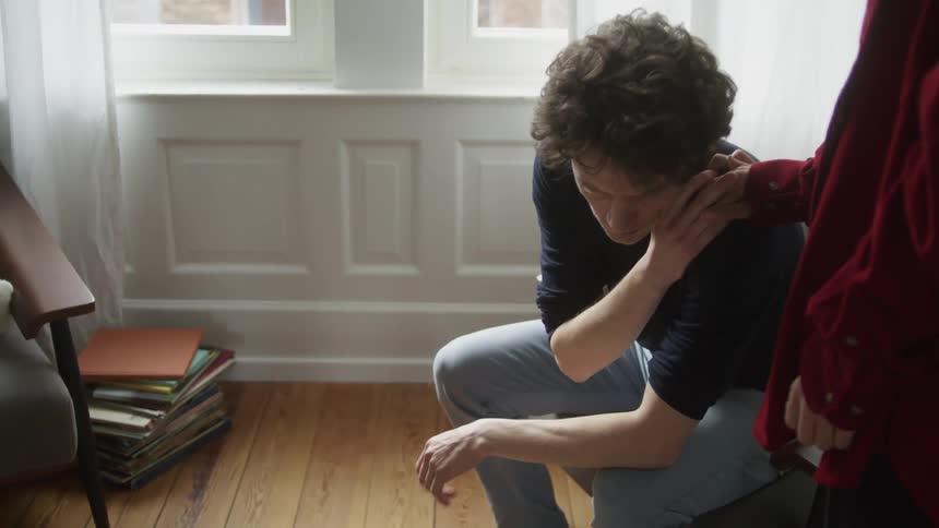 A person is consoling a sad man sitting in a chair on a room