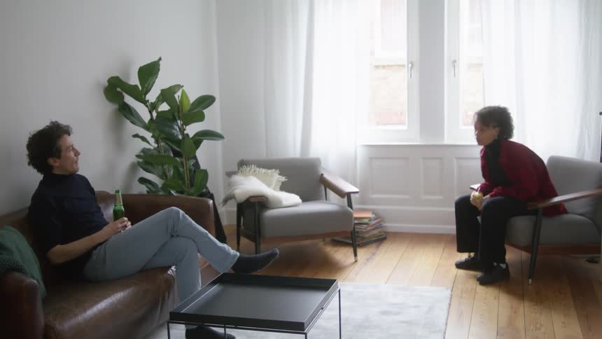 A man and a woman in a room sitting on couches with a window in the background