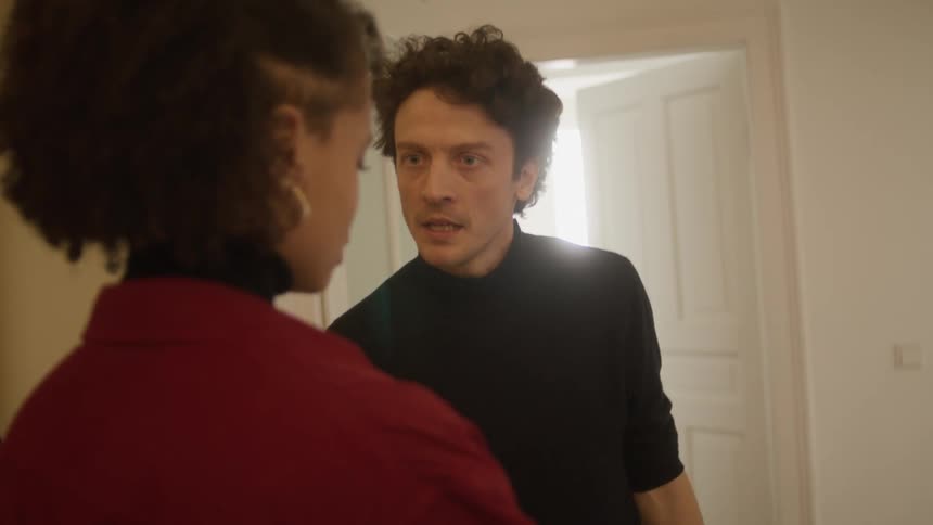 A man and a woman in a room talking with a door and a wall in the background