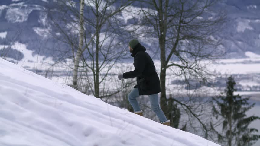 A man wearing a black parka climbing a snow covered hill near a lakeside small town in the mountains