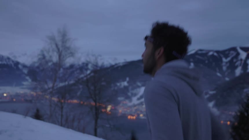 A man jogs on a snowy cliff overlooking a town along a river through a mountain valley on a cloudy evening