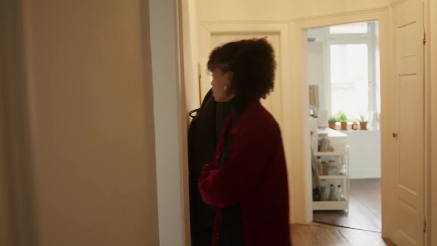 A woman with her arms folded while speaking angrily walks into a living room where a man is staring out of a window at daytime