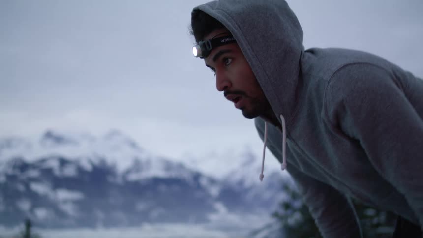A tired man wearing a sweater and a headlamp takes a breath outdoors near the snow covered mountain range