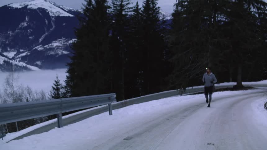 A man runs on a snowy road with tall evergreens and mountains in the distance