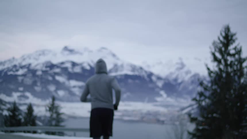 A man with a flashlight on his head with a hoodie and gloves jogging on a road with a mountain range the sky and clouds in the background