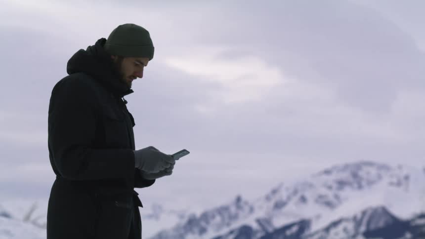Young man wearing a parka texting on his smartphone near the snow covered peaks of a mountain range