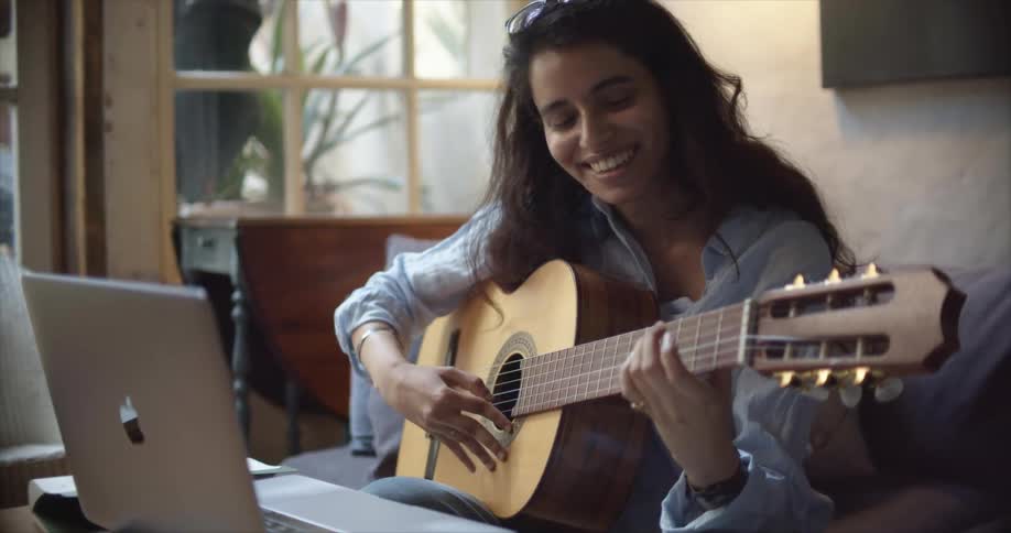 A woman talking on a video chat sitting on a sofa in a living room in front of a laptop while she plucks the string on a guitar she is holding and smiles in