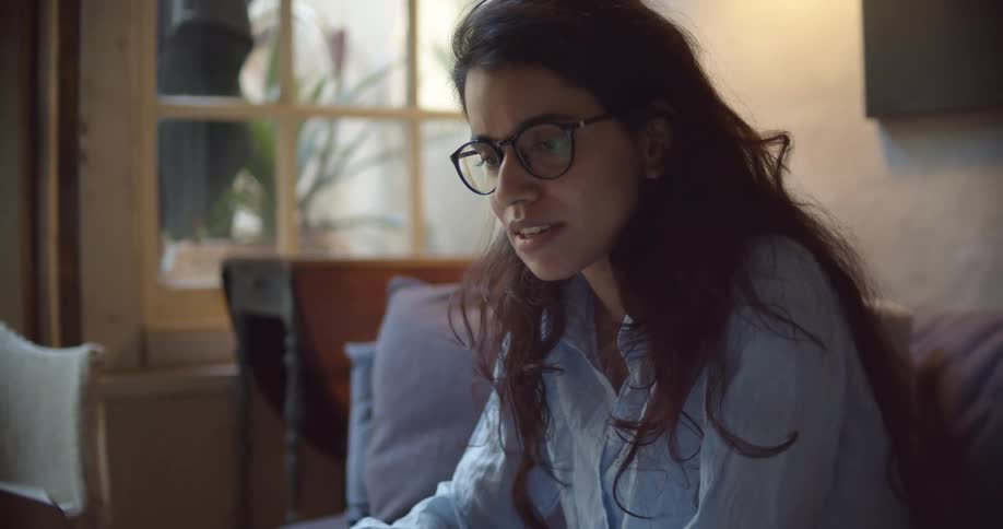 A woman at home sits on her couch and writes as she talks with someone on her laptop