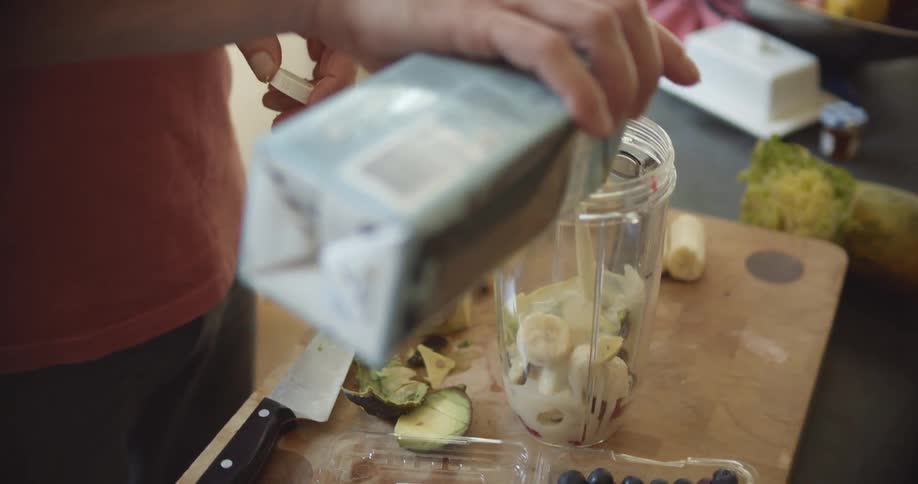 A man shakes up a bottle of plant milk and pours it into a blender on a countertop in a kitchen at daytime