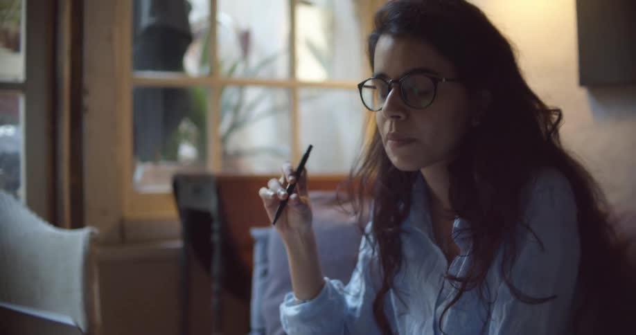 A woman with glasses sitting on a couch writing on a book with a pen while looking at a laptop