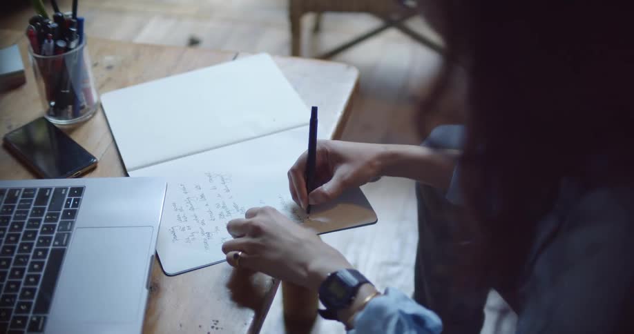 A woman in a room sitting by a table with a laptop writing on a book with a pen