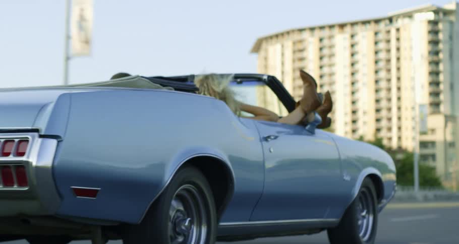Woman with her feet hanging out a convertible car driving on the highway through the city at morning