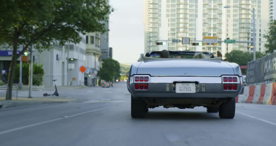 A man and a woman in a convertible on a city street with buildings the sky and clouds in the background