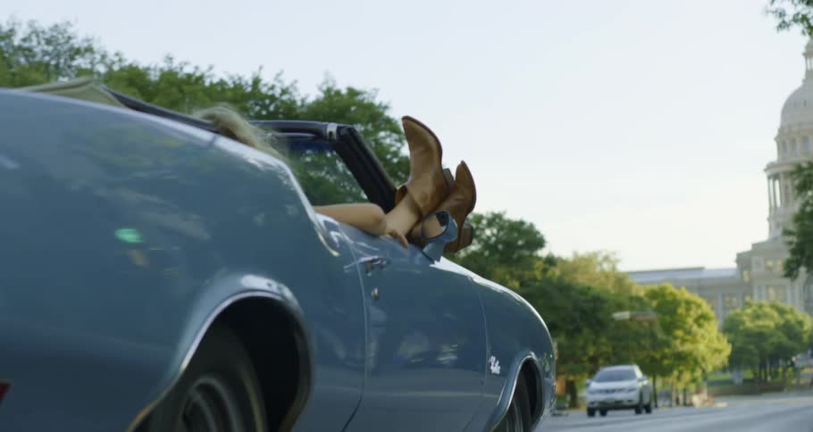 A woman relaxes with her legs up in the passenger seat of a car as it travels down a road towards a government building