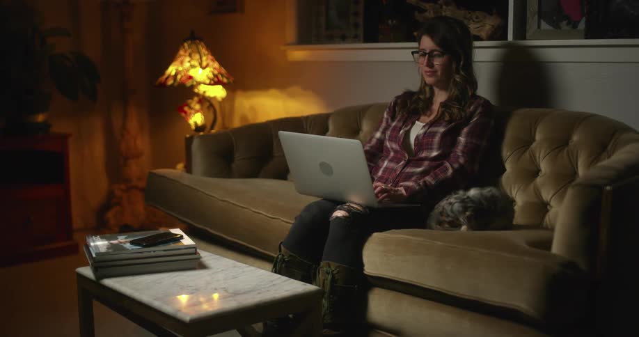 A woman sits in a living room on a couch typing on a laptop on her lap then closes it and places it on a coffee table in front of her