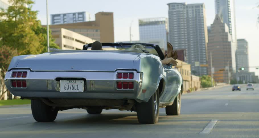 A man drives a silver convertible with a texas license plate down a street in downtown while a woman in the passenger seat sits with her feet out of the car