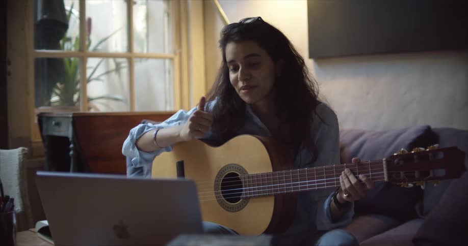 A woman sits at home in front of her computer while holding a guitar and waves at the screen and talks to someone as they teach her how to play guitar