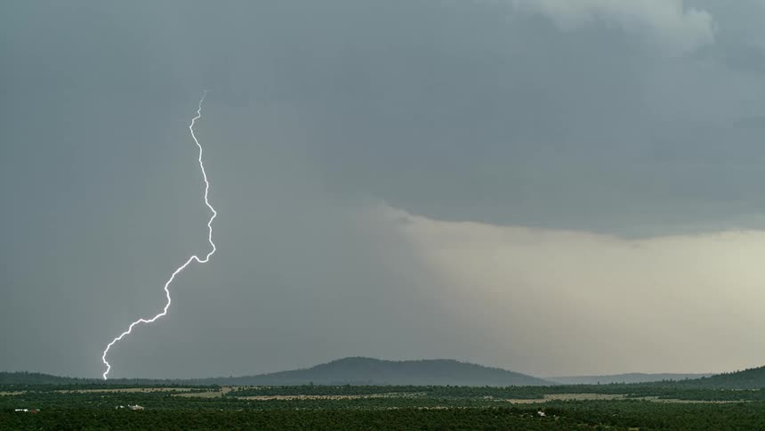 Lightning strikes down from storm clouds in the sky towards the ground with mountains in the distance at daytime