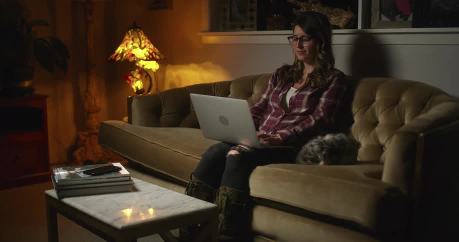 A woman sits on a couch typing on a laptop in a dimly light room