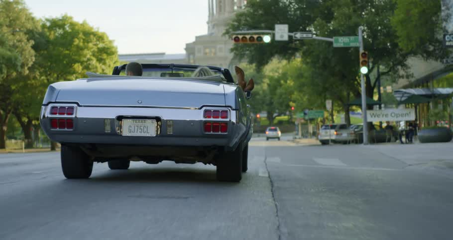 A man driving a silver convertible down a street in a city with a female passenger who has her feet propped up outside of the car at daytime