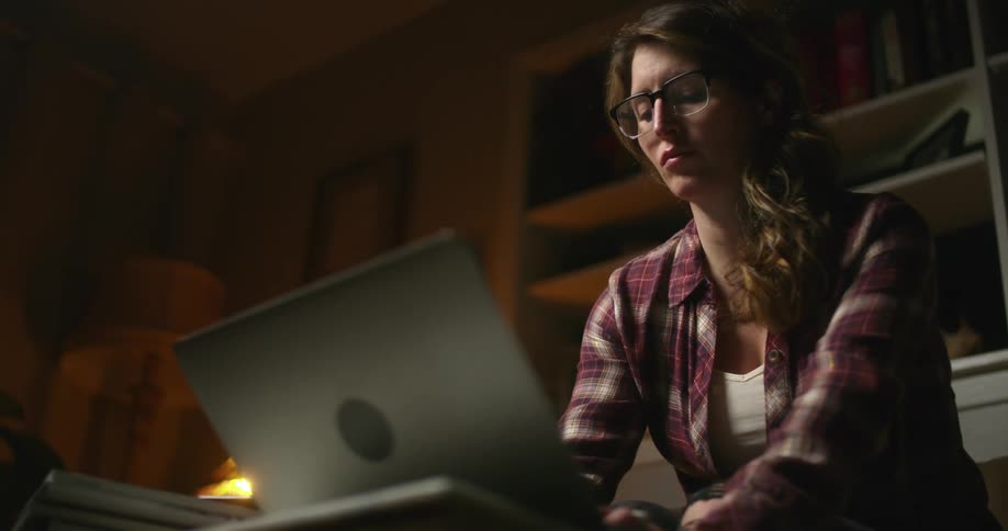 A woman in a room with glasses sitting and typing on a laptop