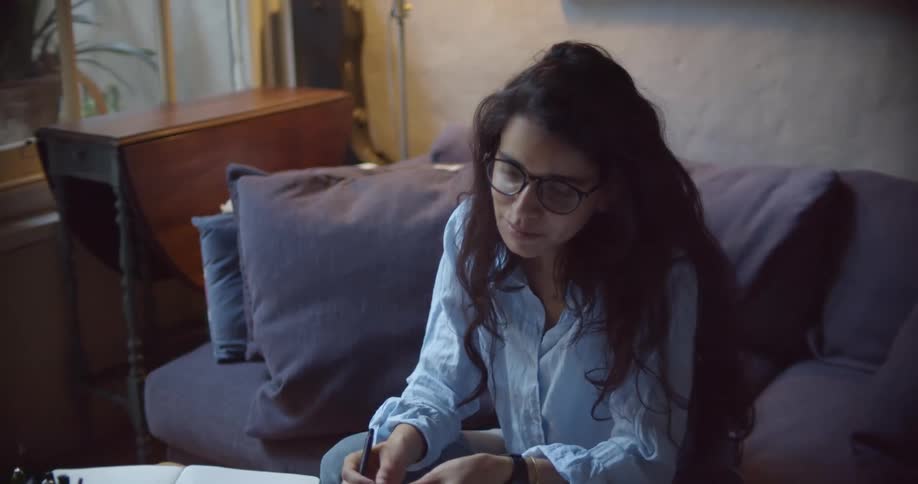 A woman sits on a sofa and writes in a notebook while talking to someone on a device and smiles