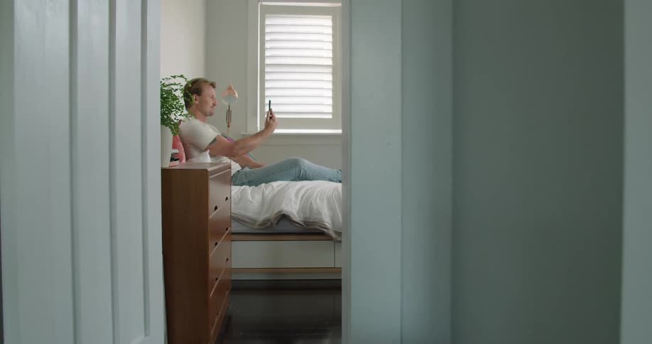 A man sits on his bed and holds his cell phone and video chats with someone