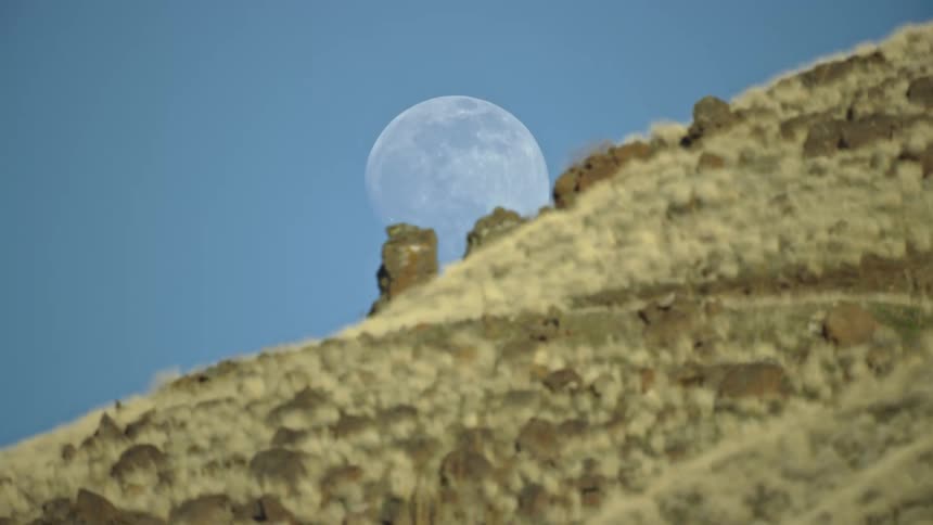 A full moon in a crystal clear blue sky in front of basalt rocks and spires on a ridge as the ridge slowly overcomes the moon in this dynamic aerial shot