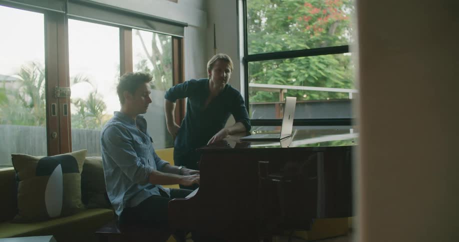A man sits at a grand piano indoors while another man stands next to him as they scroll on a laptop